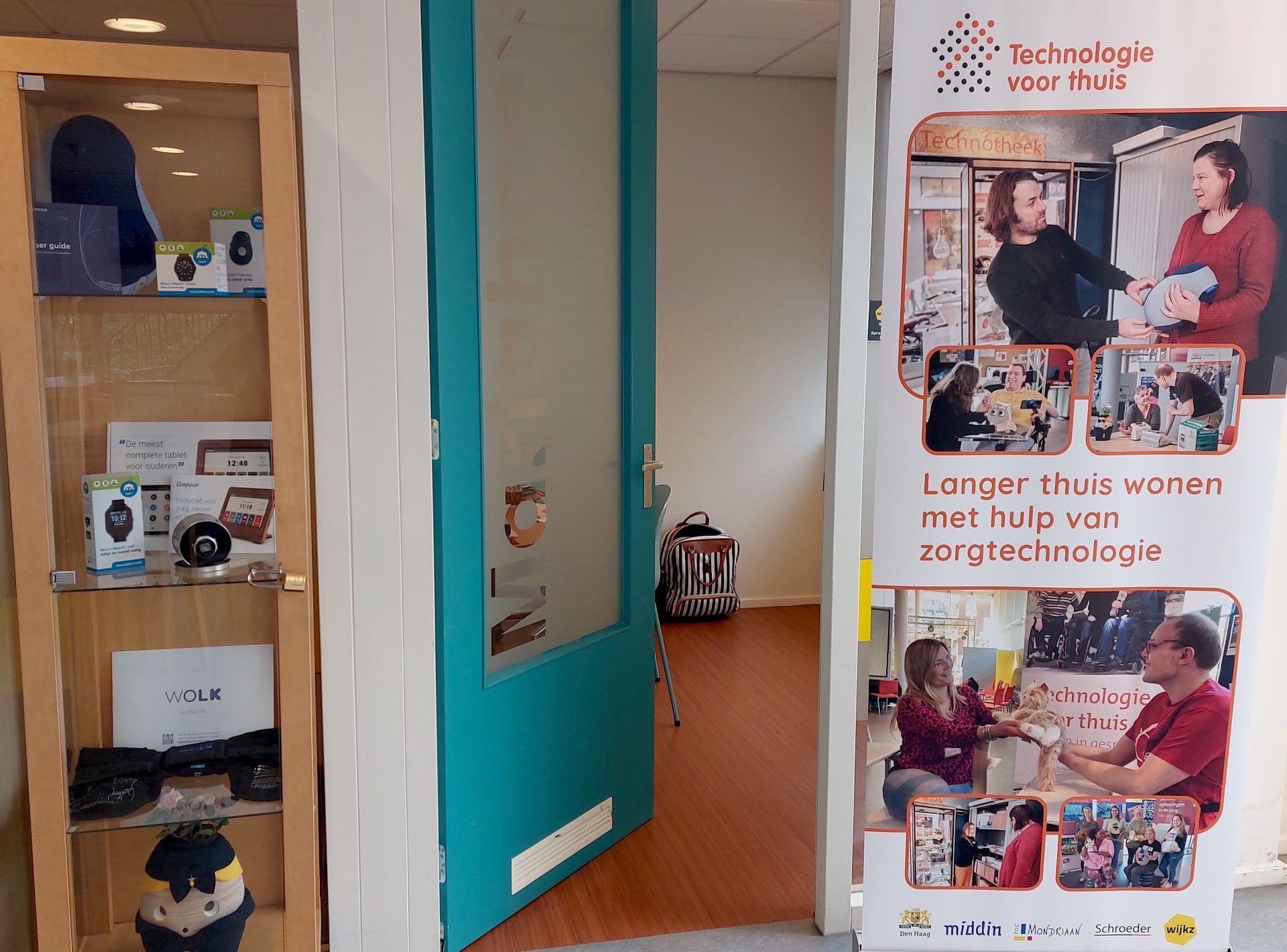 Ruimte met een vitrine met daarin verschillende hulpmiddelen en een roll-up banner met foto’s van gebruikers van zorgtechnologie