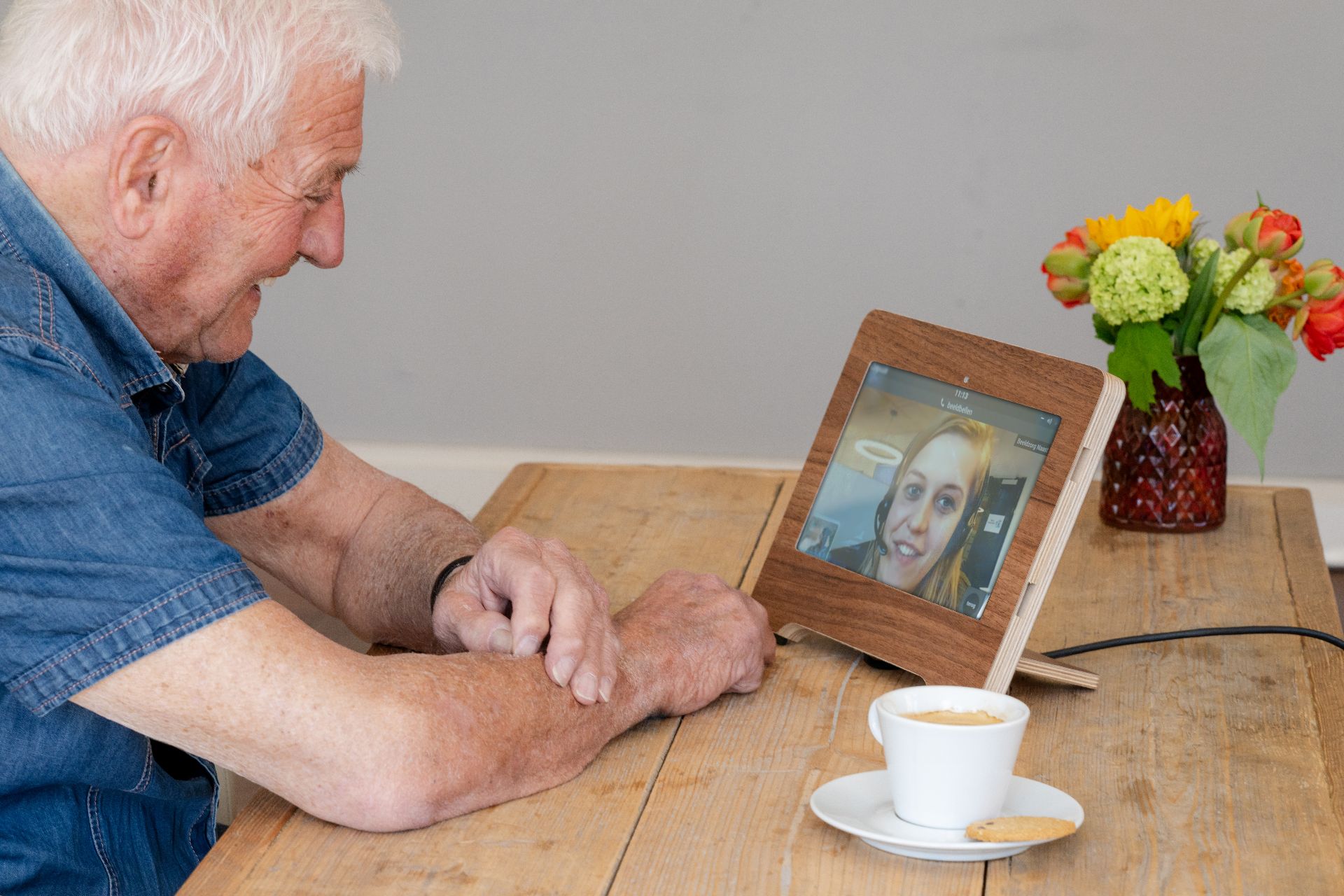 Man met grijs haar aan tafel voor een tablet met op het scherm een blonde vrouw met een koptelefoon op