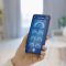 Hand in een woonkamer die een mobiele telefoon vasthoudt met op het scherm de tekst ‘smart home’ en daaronder 7 knoppen Hand in een woonkamer die een mobiele telefoon vasthoudt met op het scherm de tekst ‘smart home’ en daaronder 7 knoppen
