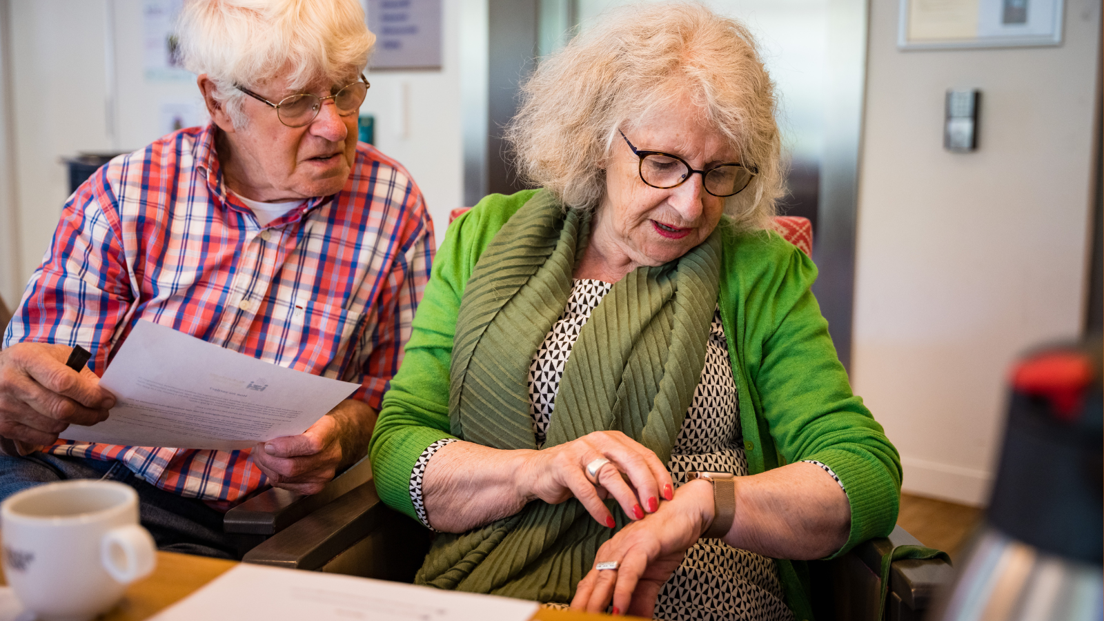 Een oudere man en vrouw met grijs haren kijken naar een alarmhorloge om de pols van de vrouw. De man heeft een informatieflyer vast